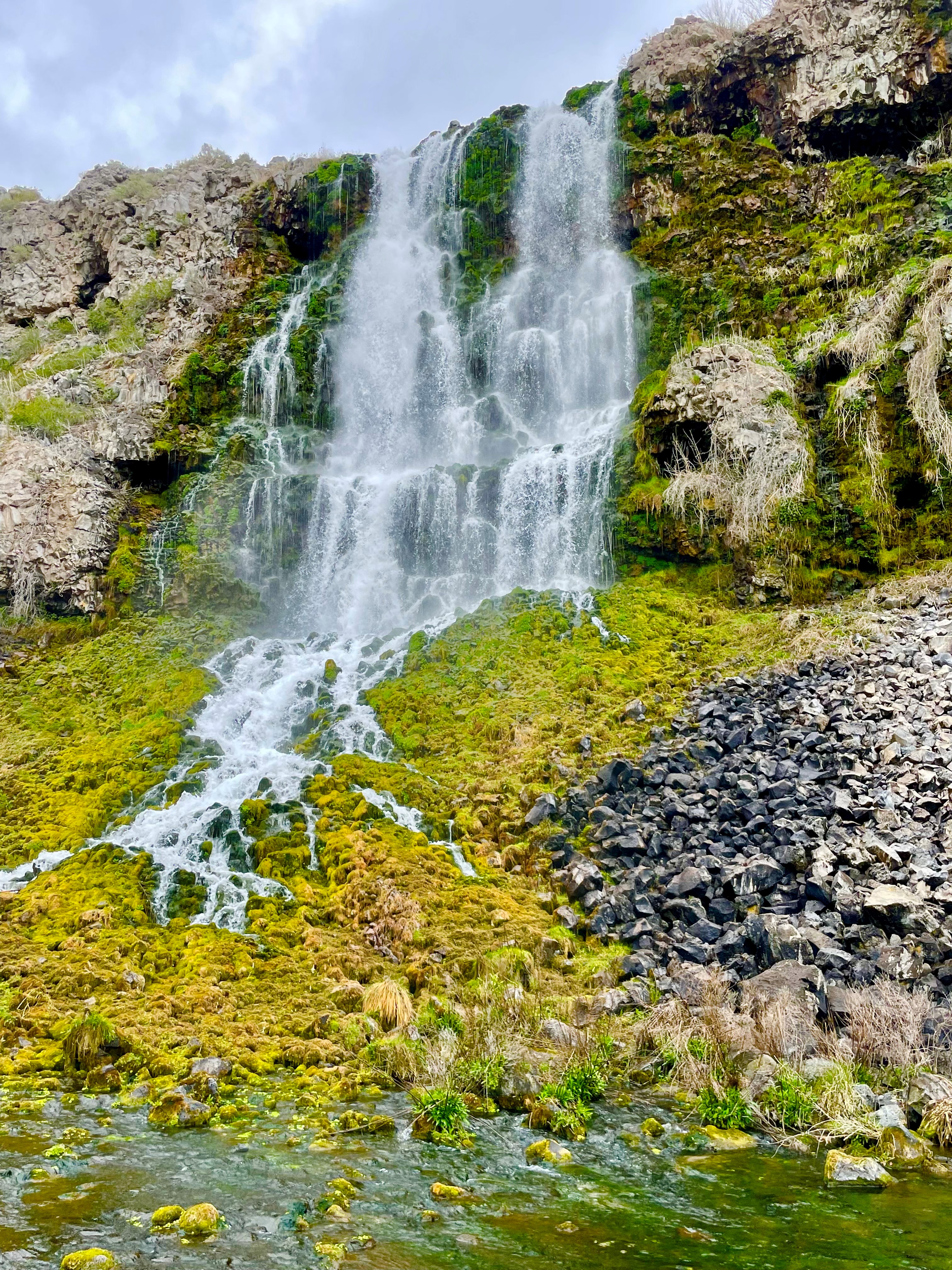 View of a waterfall, where the water cascades from a cliff on to lime green algae and moss.